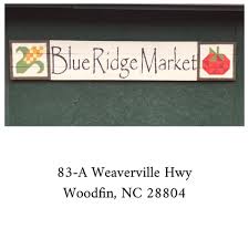 On wooden signs on a green background sit an ear of corn, Blue Ridge Market, and an apple. At the bottom reads text, 83-A Weaverville Hwy, Woodfin, NC 28804